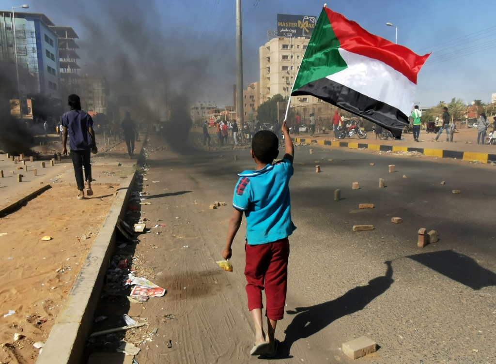 طفل يلوح بالعلم الوطني السوداني بينما يغلق محتجون شارعًا في العاصمة الخرطوم ، خلال مظاهرة ضد مقتل العشرات في حملة قمع منذ الانقلاب العسكري العام الماضي ، في 20 يناير / كانون الثاني 2022 (أ ف ب)