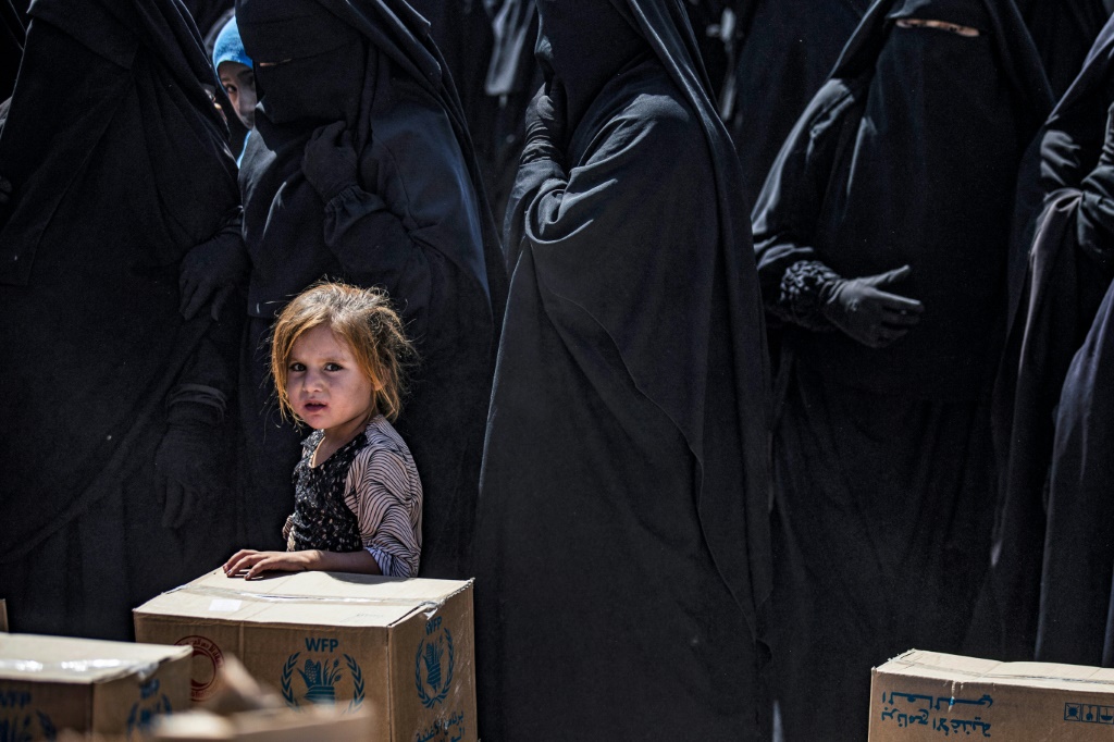     نساء وطفل يصطفون لتلقي طرود المساعدات الإنسانية في مخيم الهول الذي يديره الأكراد في سوريا (أ ف ب)