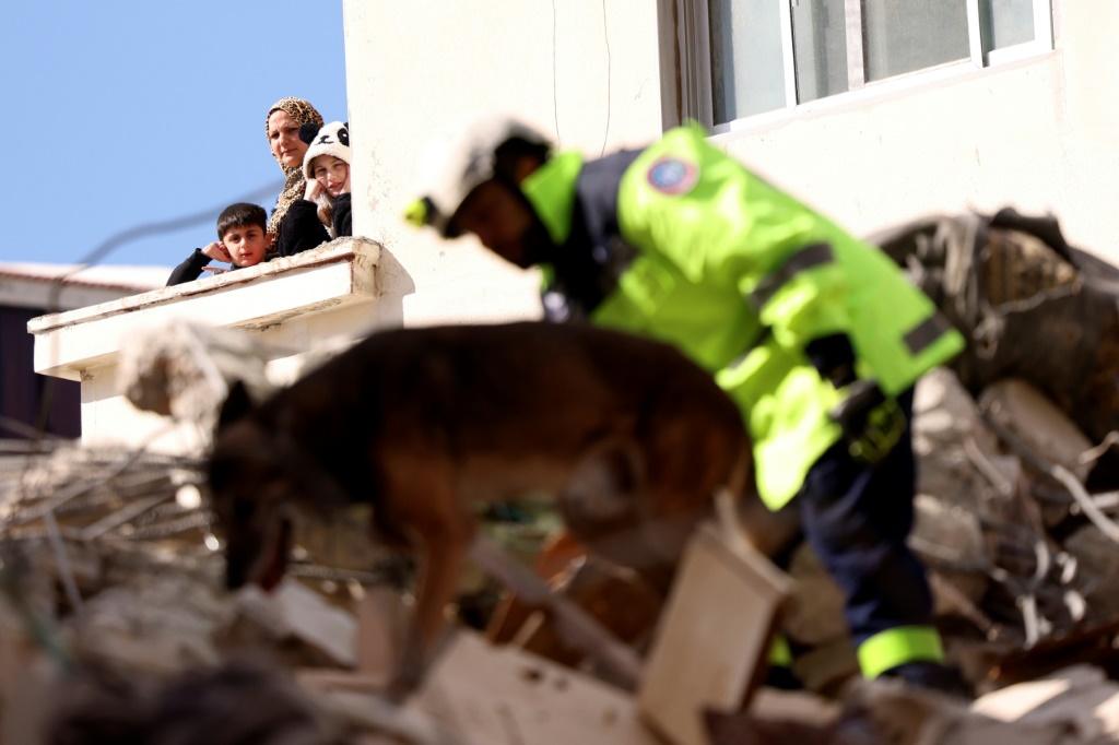 سكان يشاهدون عملية بحث يجريها عنصر من فرقة بحث وإنقاذ إماراتية تابعة لشرطة أبو ظبي برفقة كلب مدرّب في 10 شباط/فبراير 2023 في مدينة جبلة الساحلية المتضررة بشدة جراء الزلزال المدمر الذي ضرب سوريا ومقرّه تركيا متسبباً بمقتل أكثر من 28 ألف شخص في البلدين (ا ف ب)