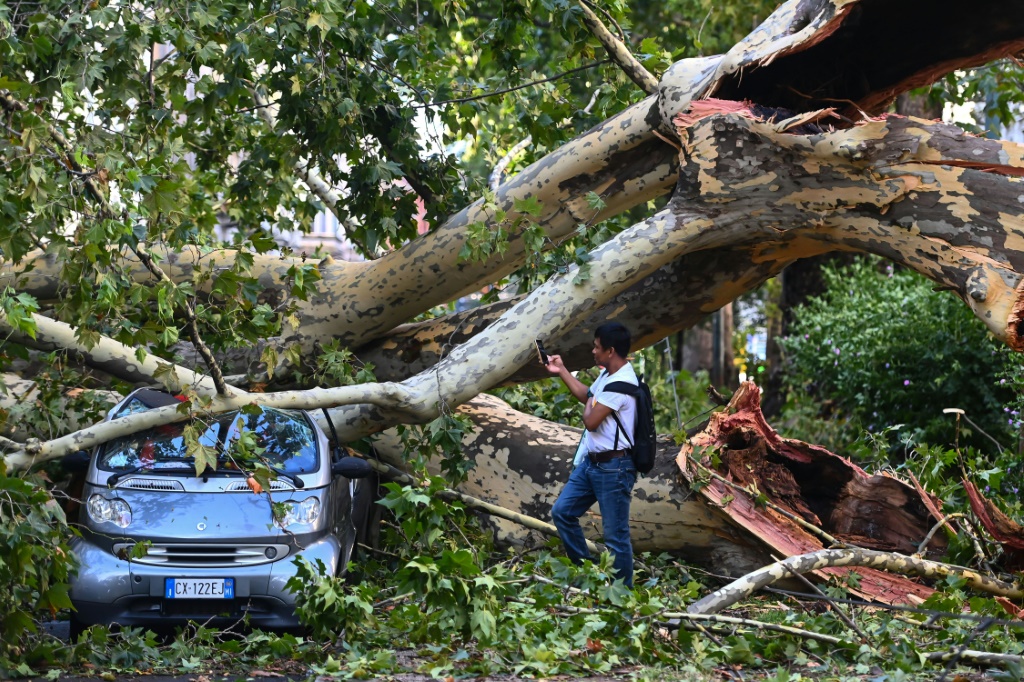     رجل يلتقط صورة لسيارة محطمة تحت شجرة سقطت في ميلانو في 25 يوليو 2023 بعد عاصفة مطرية ضربت المدينة ليلاً. (أ ف ب)   