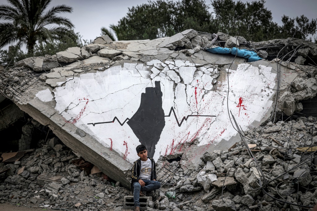 طفل فلسطيني يجلس أمام جدارية ضمن معرض أقيم على أنقاض المنازل التي دمرتها غارات جوية إسرائيلية خلال أيار/مايو 2023 في دير البلح وسط قطاع غزة في الثامن من حزيران/يونيو 2023 (ا ف ب)
