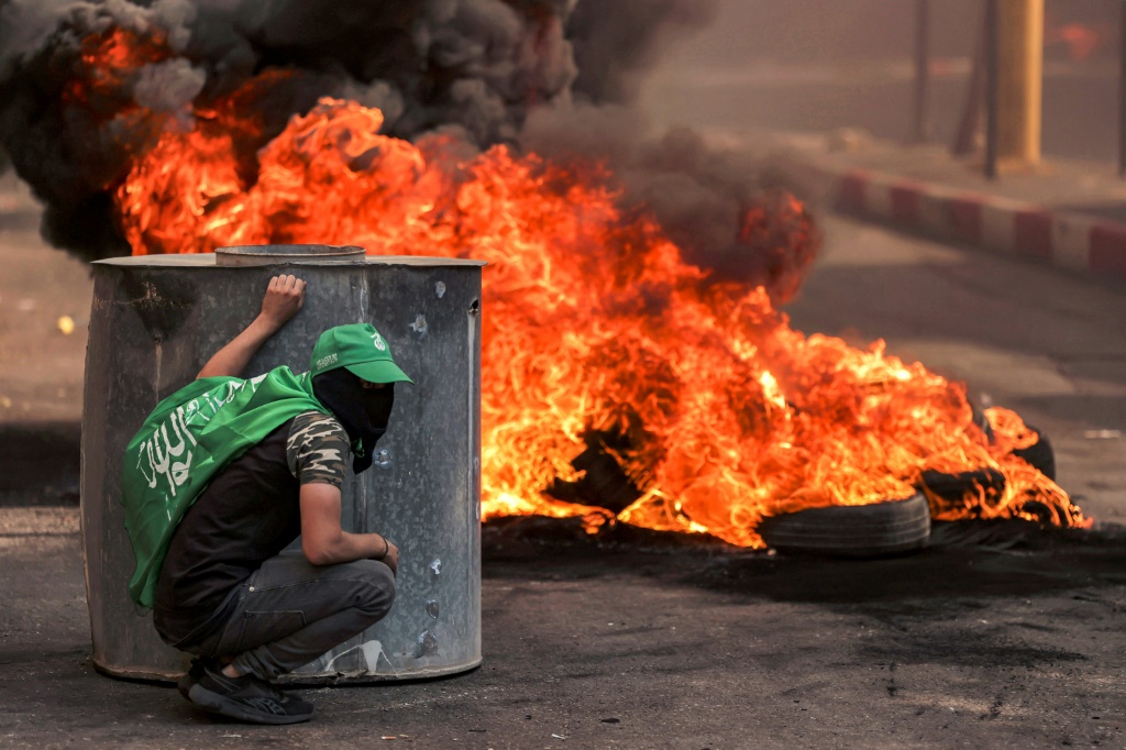 متظاهر فلسطيني ملثم يختبئ بالقرب من إطارات مشتعلة خلال اشتباكات مع القوات الإسرائيلية في أعقاب مسيرة تضامنية مع غزة نظمها أنصار حركتي فتح وحماس، في مدينة الخليل بالضفة الغربية المحتلة في 13 تشرين الأول/أكتوبر 2023 (أ ف ب)  &nbsp;