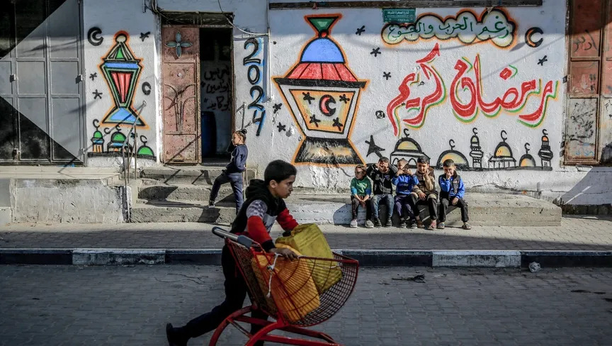 طفل فلسطيني يدفع عربة تحمل عبوات مياه أمام جدار يحمل رسومات رمضانية وسط حرب إسرائيلية مستمرة منذ 5 أشهر، قطاع غزة. 8 مارس 2024 (أ ف ب)