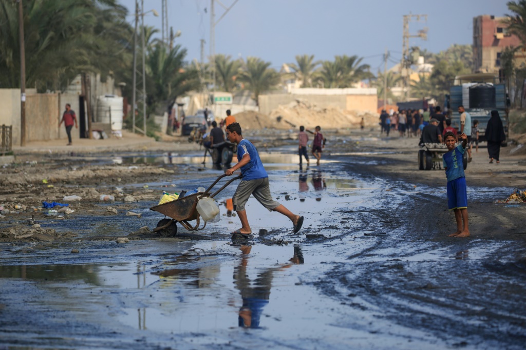 فلسطينيون يعبرون شارعًا غارقًا بمياه الصرف الصحي في دير البلح وسط قطاع غزة في 23 تموز/يوليو 2024، بعد تدمير القصف الإسرائيلي للبنية التحتية في القطاع المحاصر (أ ف ب)   