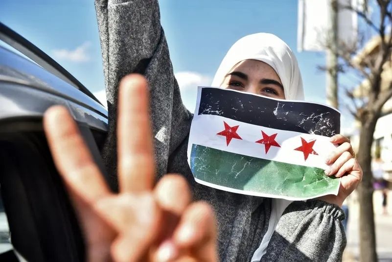 مؤيدون وأفراد من المجتمع السوري يلوحون بعلم المعارضة السورية، المعروف أيضًا باسم علم الثورة، ويرفعون لافتة تصور الرئيس السوري المخلوع بشار الأسد بعينين متقاطعتين، خلال تجمع في ساحة ترافالغار، وسط لندن، في 8 ديسمبر 2024. (أ ف ب)