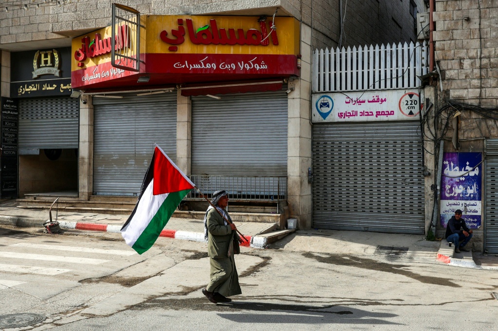 رجل يمشي أمام متجر مغلق يحمل علما فلسطينيا أثناء إضراب عام في مدينة جنين بالضفة الغربية المحتلة في 10 كانون الأول/ديسمبر 2024، بعد يوم من الاشتباكات بين قوات الأمن الفلسطينية ومقاتلين (ا ف ب)   