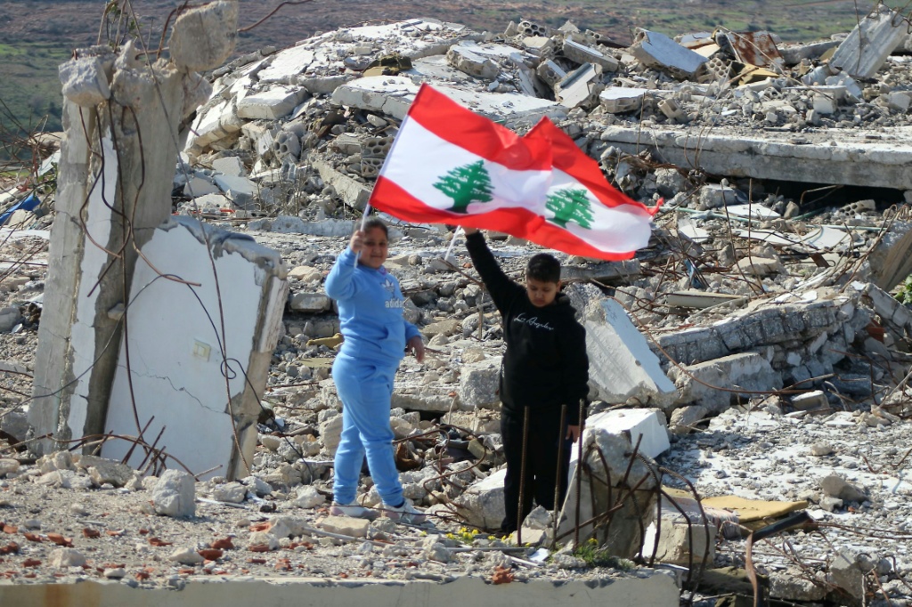 طفلان يحملان العلم اللبناني أثناء سيرهم فوق أنقاض مبنى مدمر في قرية مروحين الجنوبية، بالقرب من السياج الحدودي مع إسرائيل في 28 كانون الثاني/يناير 2025 (أ ف ب)   