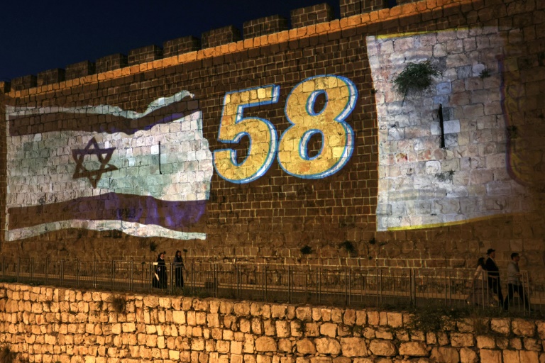 أناس يسيرون بمحاذاة أسوار البلدة القديمة في القدس في 25 أيار/مايو 2025، والتي أضيئت بالعلم الإسرائيلي ورقم 58 الذي يشير إلى ذكرى توحيد القدس واحتلالها في العام 1967(أ ف ب)
