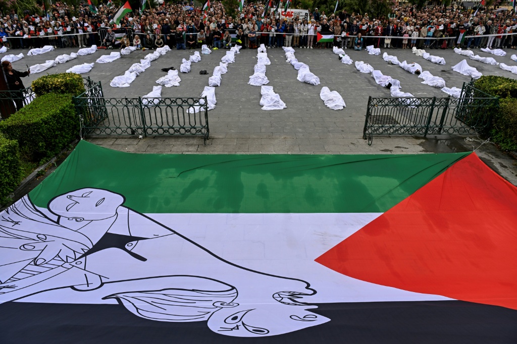 علم فلسطيني وصورة من لوحة بابلو بيكاسو "غيرنيكا" لأم تحمل طفلها، إلى جانب متظاهرين مستلقين على الأرض داخل أكفان، ضمن فعالية احتجاجية تضامنا مع الشعب الفلسطيني في غزة، تحت شعار "أوقفوا الإبادة الجماعية، اقطعوا العلاقات مع إسرائيل الآن!" في مدينة سان سيباستيان الإسبانية الباسكية، في الأول من حزيران/يونيو 2025. (أ ف ب)