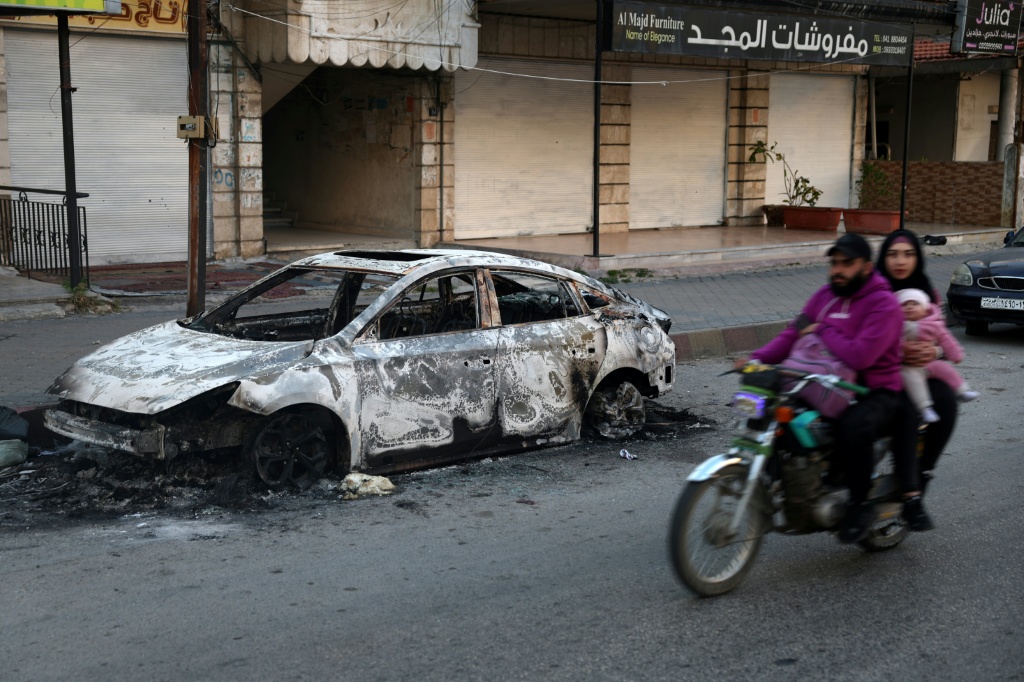 عائلة على متن دراجة نارية تمر من أمام سيارة متفحمة في جبلة في محافظة اللاذقية في الساحل السوري بتاريخ 12 آذار/مارس 2025 (أ ف ب)