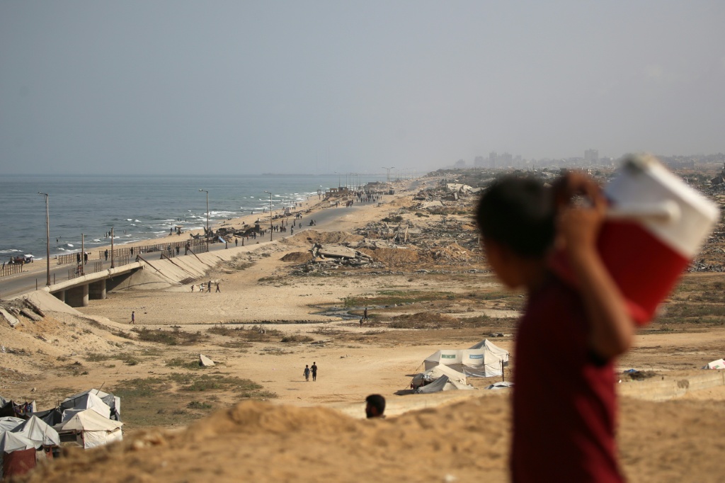 فتى فلسطيني يحمل وعاءً من الماء وهو ينظر إلى الناس يسيرون على طريق ساحلي شمال غرب مخيم النصيرات للاجئين بعد نزوحهم جنوبا في أعقاب إعلان إسرائيلي عن إغلاق طريق الرشيد باتجاه شمال قطاع غزة المحاصر في 4 تشرين الأول/أكتوبر 2025 (أ ف ب)