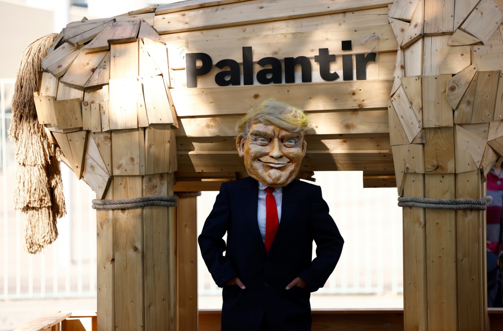 ناشط من مجموعة كامباكت! يضع رأسا يشبه الرئيس الأميركي دونالد ترامب، يقف بجانب حصان خشبي يرمز إلى حصان طروادة ويحمل اسم شركة بالانتير الأميركية في برلين في 3 أيلول/سبتمبر 2025 (ا ف ب)