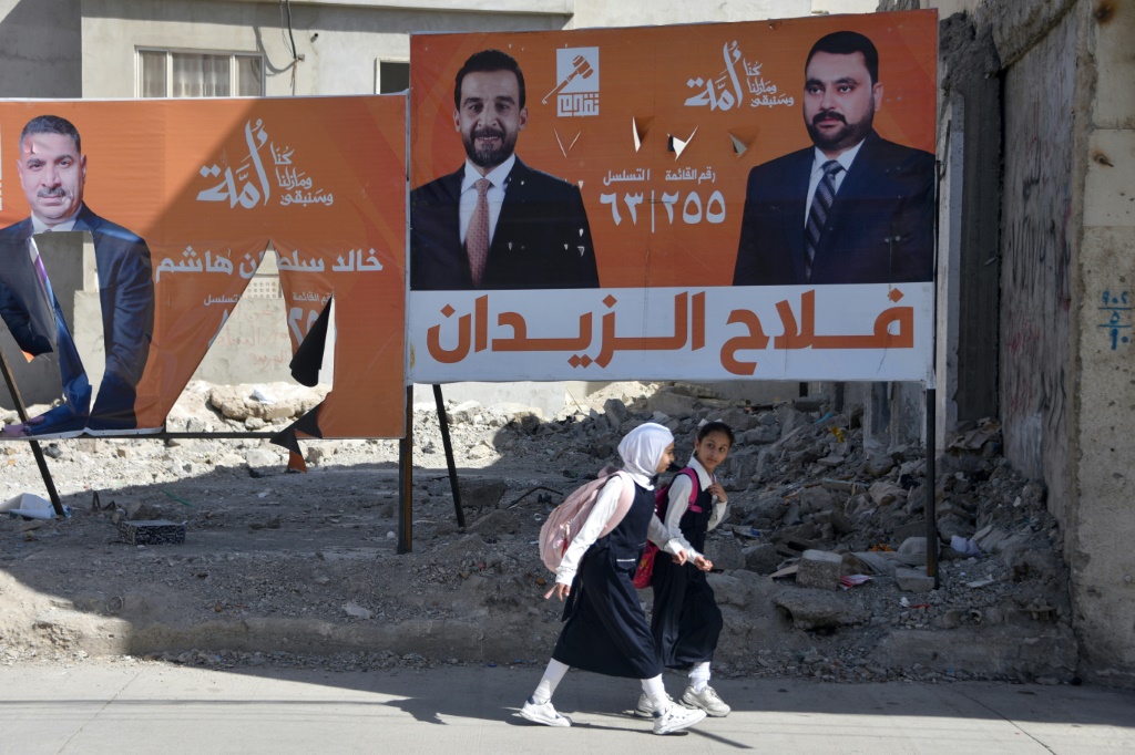 فتاتان تسيران إلى جانب لافتتين انتخابيتَين لمرشحَين مع قائمة حزب "تقدمّ" السني في الانتخابات التشريعية العراقية، في 28 تشرين الأول/أكتوبر 2025 في الموصل بشمال العراق (ا ف ب)