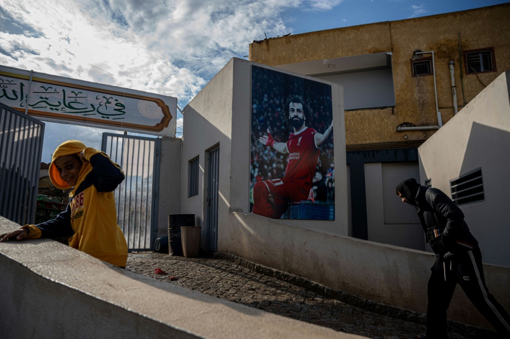يمرّ مصريون أمام صورة لاعب ليفربول محمد صلاح في مركزه المخصص للشباب والواقع في قرية نجريج المصرية، على بعد نحو 120 كيلومتراً شمال غرب القاهرة، في 10 كانون الأول/ديسمبر 2025 (ا ف ب)