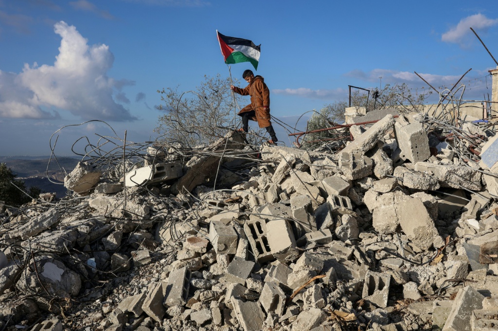 طفل يرفع العلم الفلسطيني فوق أنقاض منزل عائلة مالك سالم الذي هدمته آليات الجيش الإسرائيلي الذي يتهم سالم بتنفيذ هجوم إطلاق نار وطعن صيف 2025، في قرية بزاريا في شمال غرب محافظة نتابلس في شمال الضفة الغربية المحتلة في 24 كانون الاول/ديسمبر 2025 (ا ف ب)