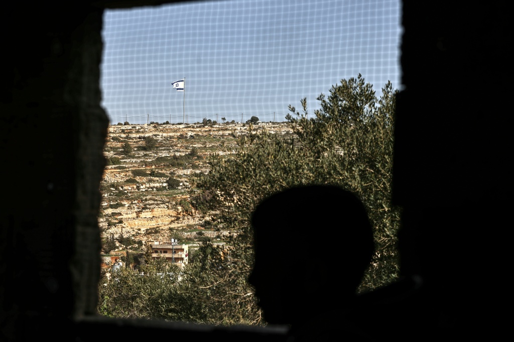 صورة التُقطت من قرية أم صفا في شمال رام الله في الضفة الغربية المحتلة، تظهر شابا فلسطينيا أمام نافذة، بينما يرفرف العلم الإسرائيلي على قمة تل يُطل على القرية، في 16 شباط/فبراير 2026 (ا ف ب)