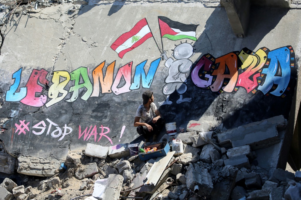 شاب فلسطيني يجلس بجوار جدارية رسمها على سطح منزل مدمر في خان يونس بجنوب قطاع غزة، لإظهار تضامنه مع شعب غزة ولبنان، في 26 أيلول/سبتمبر 2024 (ا ف ب)